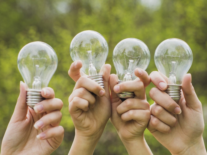 Fyra händer håller upp glödlampor utomhus med grönskande bakgrund.
