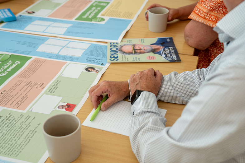 Två personer sitter vid ett bord med papper, tidning och kaffekoppar, en person skriver med en grön penna.