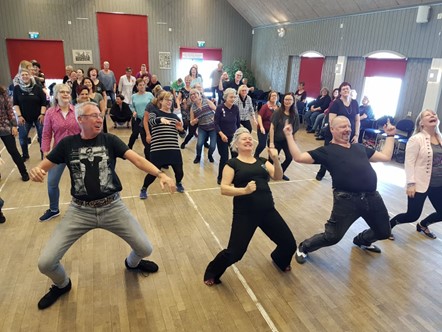 En grupp vuxna dansar energiskt tillsammans i en stor sal med trägolv och röda dörrar.