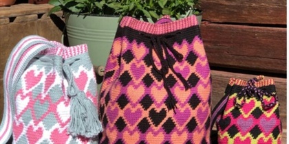 Tre färgglada virkade väskor med hjärtmönster står på en träbänk framför en blomkruka.