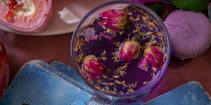 En kopp te med torkade rosor, rosa bakverk, lila macaron och blå kort med trollslända, omgivna av lila blommor.