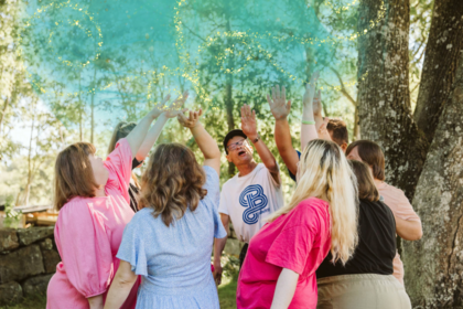 En grupp människor står i en ring utomhus och kastar färgat pulver i luften under träd.