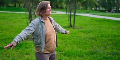 Person står med utsträckta armar i en grön park, ser glad ut och njuter av naturen.