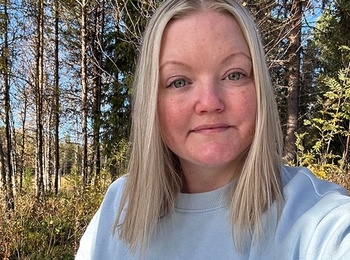 Kvinna med ljust hår och ljus tröja sitter utomhus i en solig skogsmiljö.