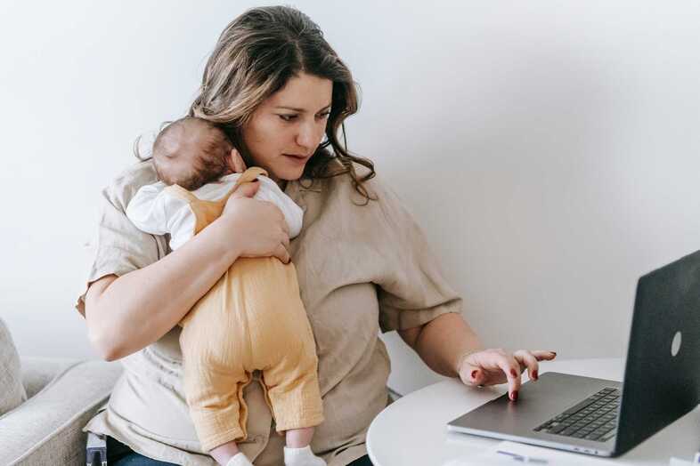 En kvinna håller en baby mot sig med ena armen och skrollar på en laptop med den andra handen.