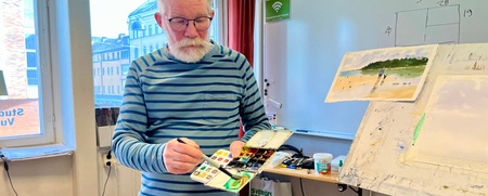 En äldre man med glasögon och randig tröja håller en färgpalett i ett ljust klassrum med staffli och målning.
