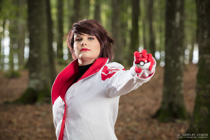 Person i vit och röd cosplaydräkt håller fram en pokéboll i en skogsmiljö.
