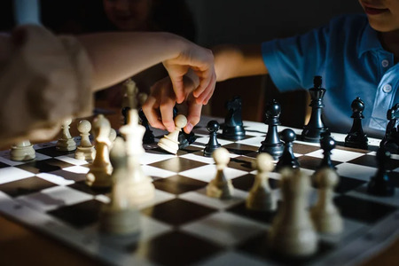 Två personer spelar schack, en hand flyttar en pjäs på brädet, starkt ljus faller över spelet.