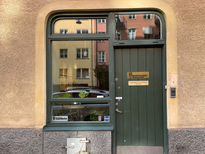 Grön dörr med glasfönster och skylt på en putsad fasad, reflektioner av hus syns i fönstret.