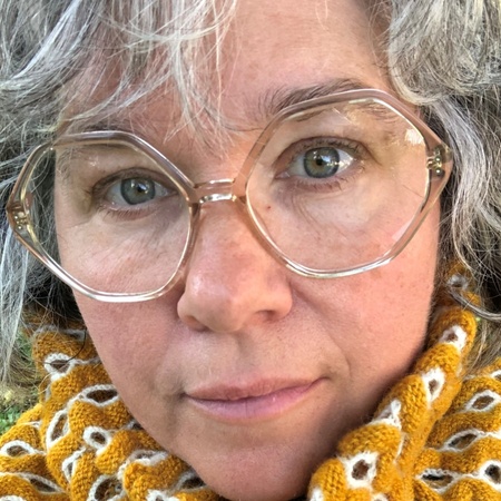Närbild på person med glasögon och grått hår, bär en gulstickad halsduk med vitt mönster.