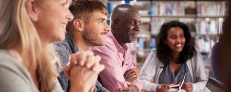 Fyra vuxna personer sitter vid ett bord i ett bibliotek och arbetar tillsammans med datorer och anteckningar.