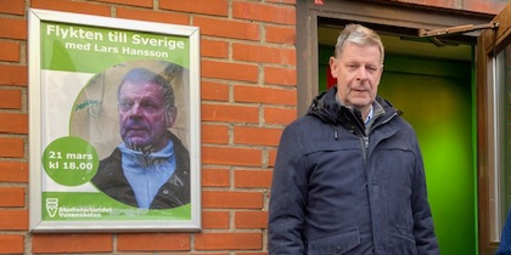 Två män står utanför en byggnad med röd tegelvägg, en affisch om ett evenemang syns på väggen.