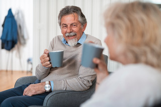 Två äldre personer sitter och dricker kaffe tillsammans i ett vardagsrum.