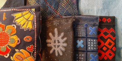 Flera textilier med färgglada broderier och mönster, inklusive kuddar och väskor, visas tillsammans.