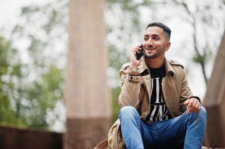 Ung man i beige trenchcoat sitter utomhus och pratar i mobiltelefon, ser glad ut.