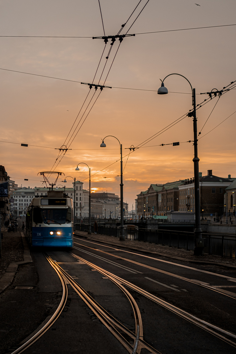 En blå spårvagn kör på spår genom en stad vid solnedgång, med gatlyktor och byggnader längs kanalen.