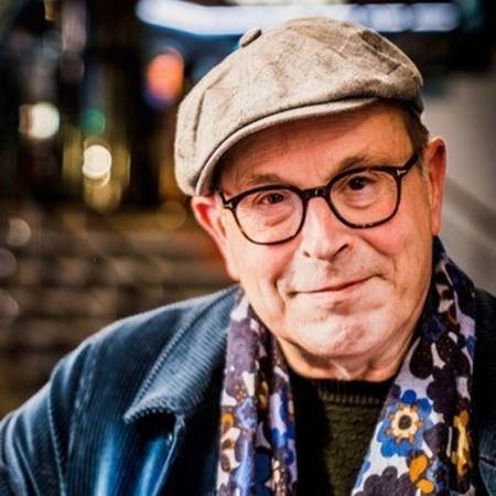 Äldre man med glasögon, keps och blommig halsduk står inomhus och ler mot kameran.