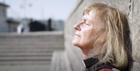 Äldre kvinna lutar sig mot en mur och blundar i solskenet utomhus.