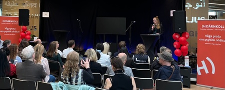 En kvinna talar på en scen inför en sittande publik i en modern lokal med text på väggen.