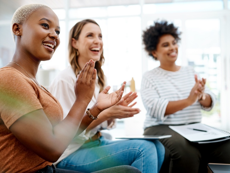 Tre kvinnor sitter tillsammans inomhus och applåderar, ser glada och engagerade ut.