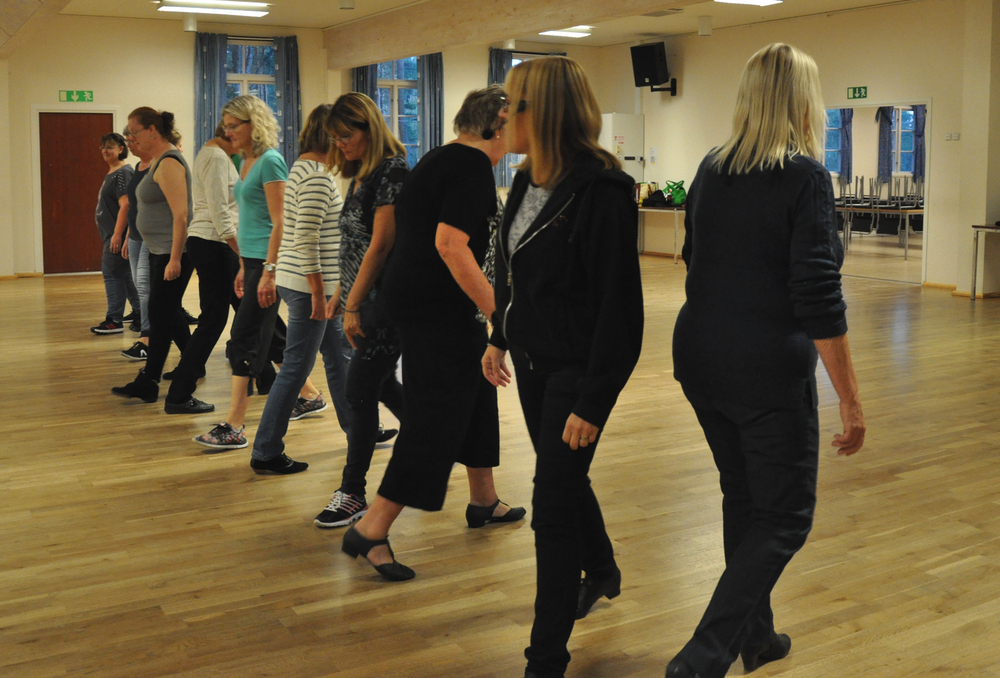 En grupp människor övar linedance i en ljus danssal med trägolv.
