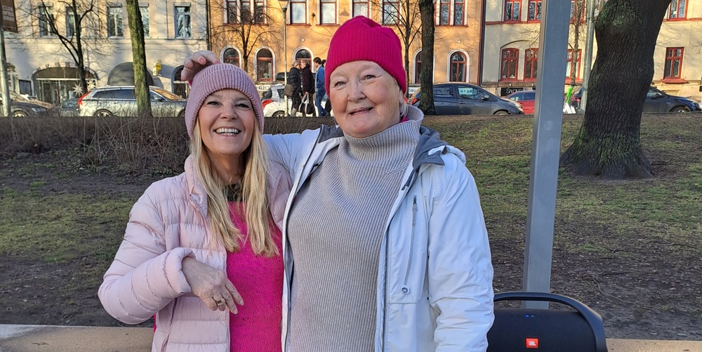 Två glada kvinnor i rosa och vita vinterkläder står utomhus i en park, båda bär mössa.