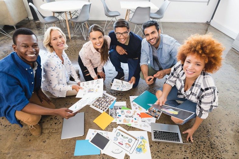 Sex personer sitter på golvet i ett kontorsrum och arbetar tillsammans med papper och datorer.