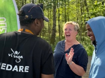 Tre personer samtalar och skrattar utomhus vid en grön flagga i en solig skogsmiljö.