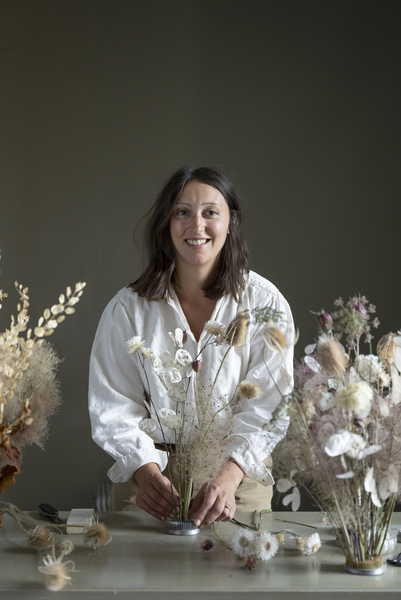 Person arrangerar torkade blommor i en vas på ett bord med fler blomsterarrangemang.