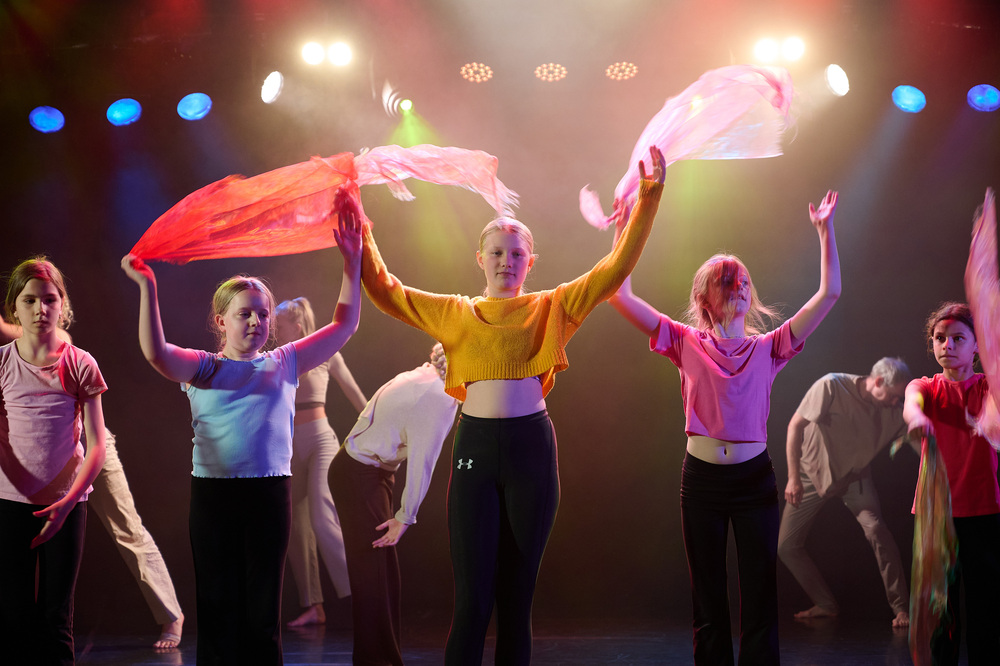 En grupp unga personer dansar på scen med färgglada sjalar under scenbelysning.