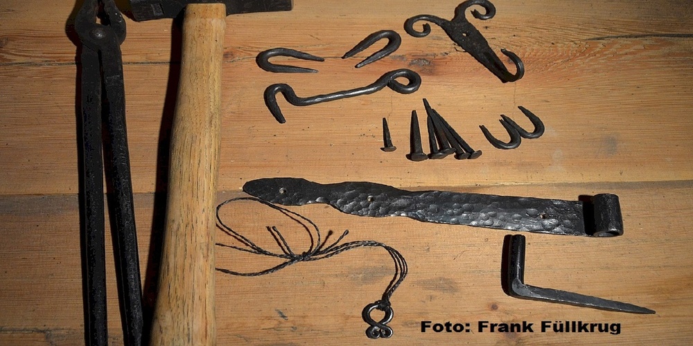 Olika smidesverktyg och smidda föremål ligger på ett träbord, inklusive hammare, tång och spikar.