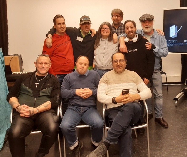 En grupp människor poserar tillsammans i ett rum med stolar, piano och en TV-skärm.