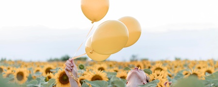 Person håller tre gula ballonger i ett fält av solrosor under en ljus himmel.