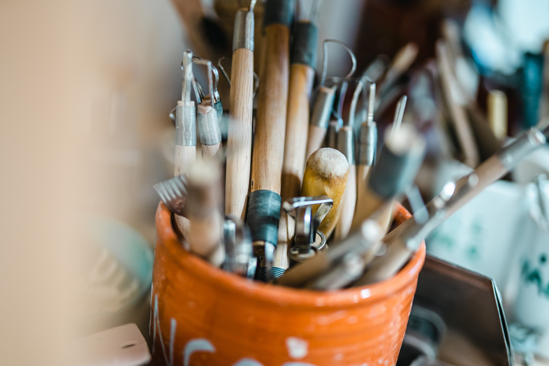 En orange kopp fylld med olika keramikverktyg i trä och metall på ett arbetsbord.