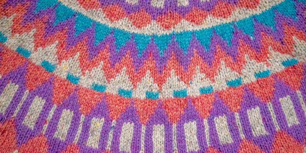 Närbild av ett stickat tyg med färgglatt geometriskt mönster i lila, turkos, rött och beige.