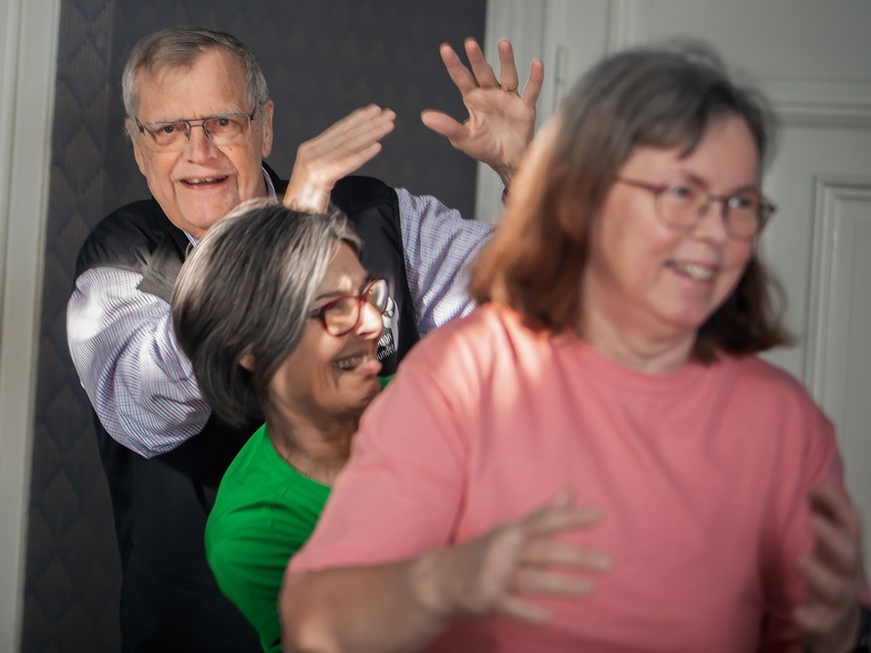 Fyra äldre personer skrattar och rör sig tillsammans inomhus, i en ljus och glad miljö.