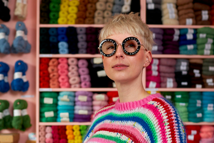 Person med kort blont hår och stora runda glasögon bär en färgglad stickad tröja i garnbutik.