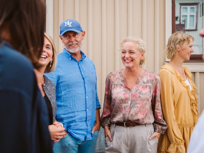En grupp vuxna står utomhus framför en beige vägg och samtalar.
