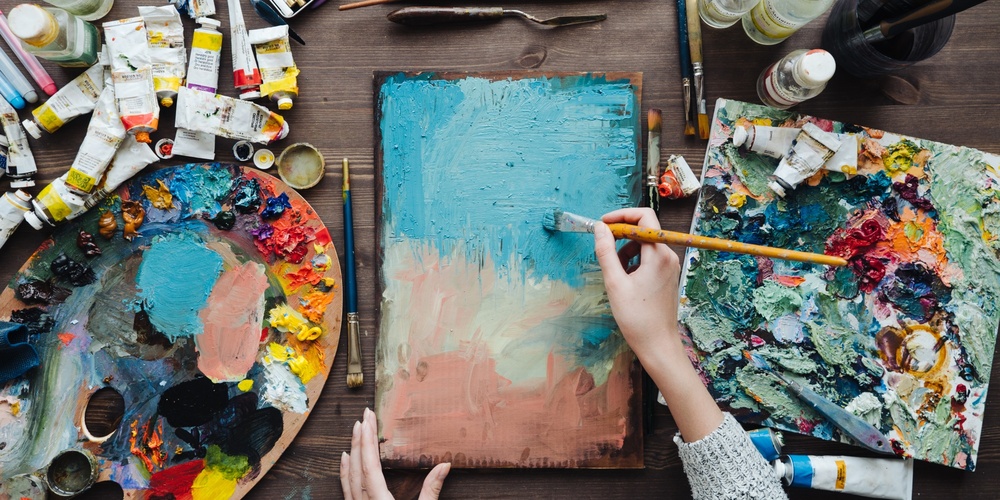 En person målar abstrakt konst på en duk med pensel, omgiven av färgtuber, penslar och färgpaletter på ett bord.