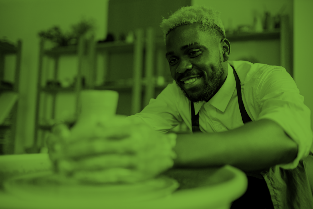 En leende person arbetar med keramik på en drejskiva i en ateljé, bilden har grön ton.