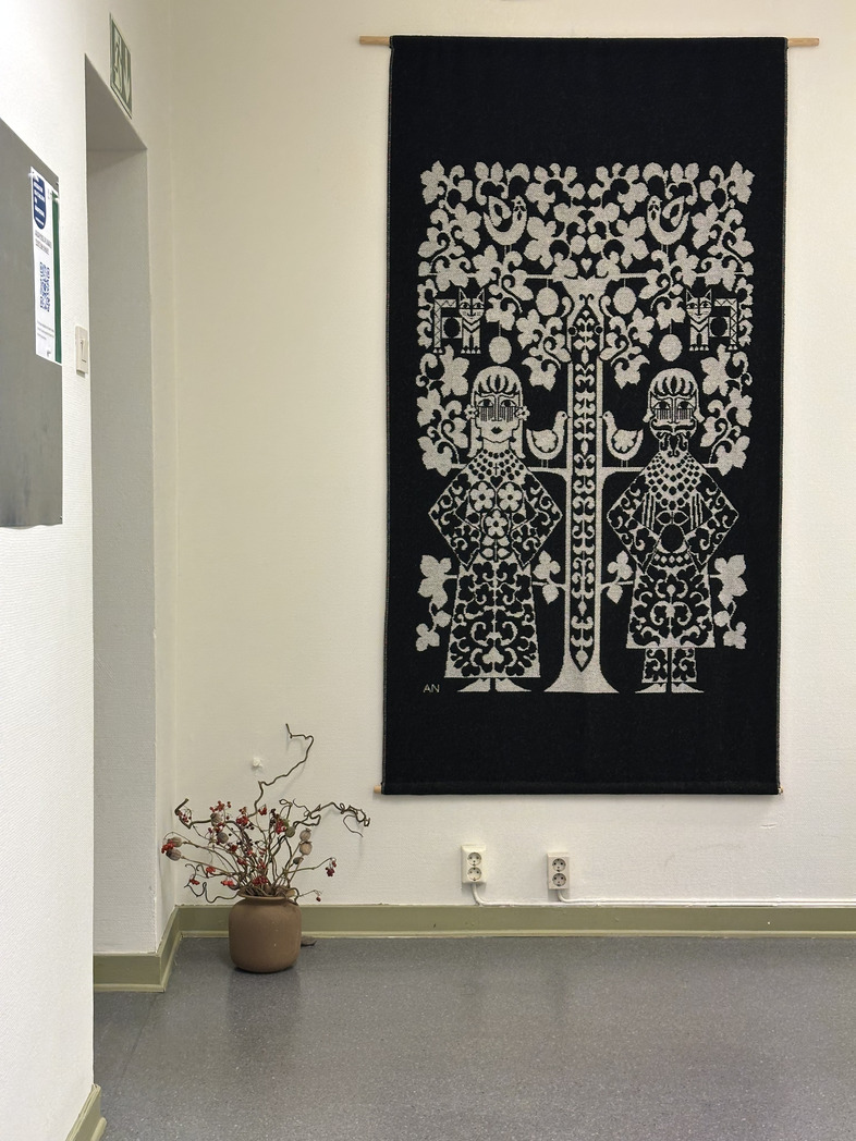 En väggbonad med två stiliserade figurer och ett träd hänger på en vit vägg ovanför ett brunt krus med torkade grenar.