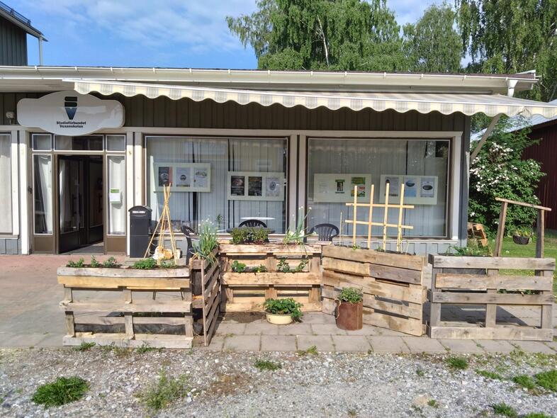 En liten butik med träpallar och växter utanför, samt stora fönster och en markis ovanför ingången.