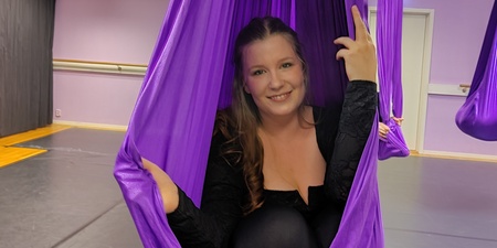 Kvinna sitter i en lila aerial yoga-hängmatta i en träningssal och ler mot kameran.