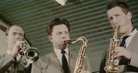 Tre män i kostym spelar blåsinstrument, två saxofoner och en trumpet, inomhus under ett tak av träpanel.