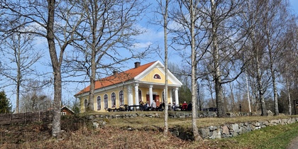 Gult hus med rött tak på en kulle, omgiven av nakna träd och människor som sitter utanför i vårsolen.