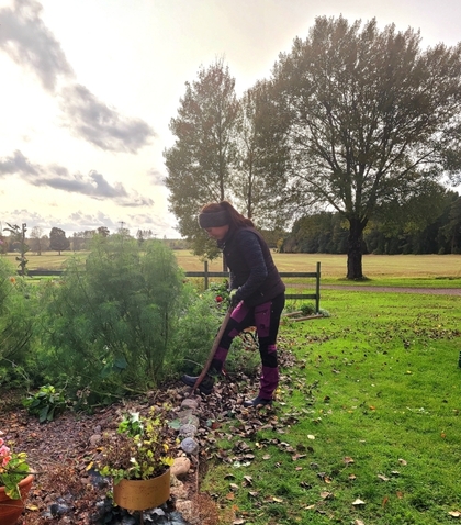 En person arbetar i en trädgård med en spade, omgiven av grönska och höstlöv under en mulen himmel.