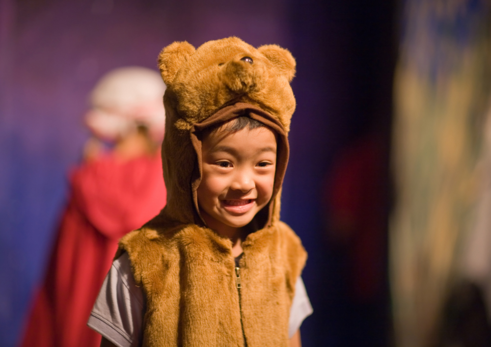 Ett barn klädd i en brun björndräkt ler på en scen med suddig bakgrund.