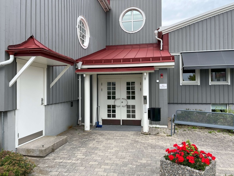 Entré till grå byggnad med röd takdetalj, vita dörrar, bänk och blomkruka med röda blommor.