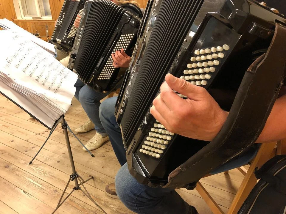 Två personer spelar dragspel vid ett notställ med noter i ett trägolv-rum.