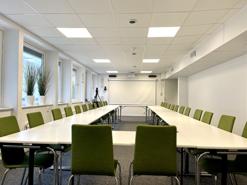 Ljust konferensrum med U-formade vita bord och gröna stolar, stora fönster och växter i fönstren.